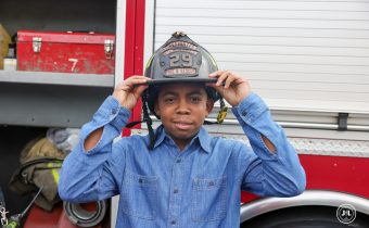 Young African-American boy wearing a denim blue shirt, standing in front of a fire truck, trying on a fire fighting helmet.
