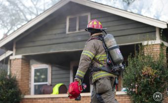 Firefighter in fire gear with air tank, walking in front of an workman's arts and crafts style bungalow, holding equipment in his hand.