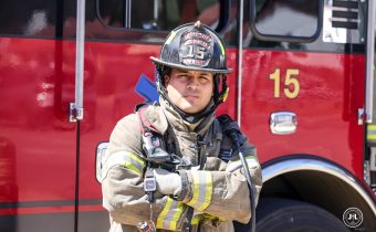 Male firefighter in full firefighting turnout gear, standing in front of a fire truck with his arms crossed.